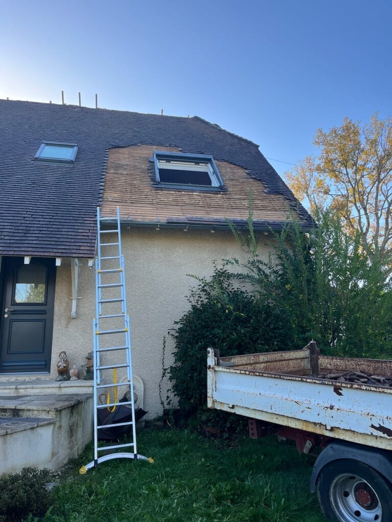 Une maison au toit pentu en cours de rénovation présente lune de nos réalisations, avec une échelle appuyée contre elle et une fenêtre ouverte. Une caravane est garée devant, et il y a de la verdure autour de la maison.