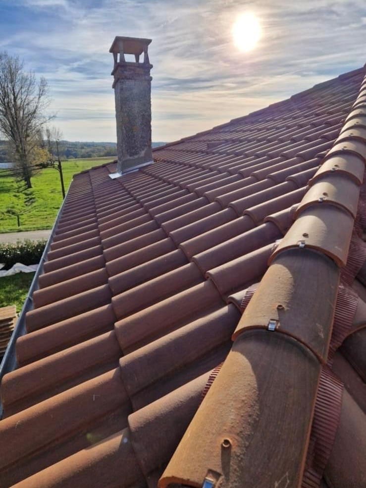 Nos réalisations présentent une scène pleine de charme : un toit de tuiles rouges avec une cheminée sous un ciel partiellement nuageux et ensoleillé. Au-delà du toit, un champ herbeux et un arbre ajoutent à la vue pittoresque.